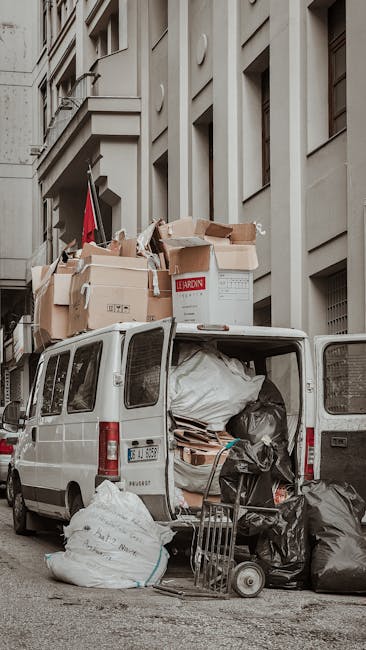 A white removal van parked on a city street in front of a multi-storey building with large windows and stone façade, with its rear doors open revealing various packing materials and household items i