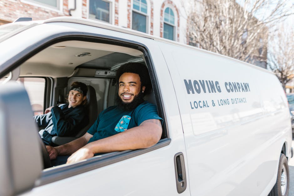 A white removal van parked on a city street in front of a multi-storey building with large windows and stone façade, with its rear doors open revealing various packing materials and household items i