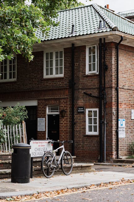 The exterior of a brick residential building with a light grey tiled roof, two small front windows, and a white front door with a black mailbox beside it. In front of the building, a bicycle is parked against a white fence, next to a black trash bin and a sign indicating parking restrictions for Chelsea Square. The pavement is partly covered with fallen leaves, and a tree with green foliage partially obscures the upper part of the building. The scene depicts a typical street view in Chelsea, suitable for home relocation or furniture transport services, with a focus on accessibility and environmental context relevant to removals companies such as Removal Van Chelsea.