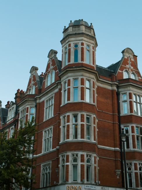 An image capturing the exterior of a historic red brick building with ornate architectural details, including large, multi-paned windows and decorative stonework. The building features a prominent turret with a rounded top and a small balcony, set against a clear blue sky. Surrounding the base are a few trees and a traditional black lamppost. The scene is taken during daylight, highlighting the building's classic Victorian or Edwardian style typical of Chelsea, London. This historic architecture can be relevant to house removals, as clients may require furniture transport and packing for relocating within or to this area, where properties often feature ornate facades and require careful handling during the loading process, which Removal Van Chelsea manages for their customers.