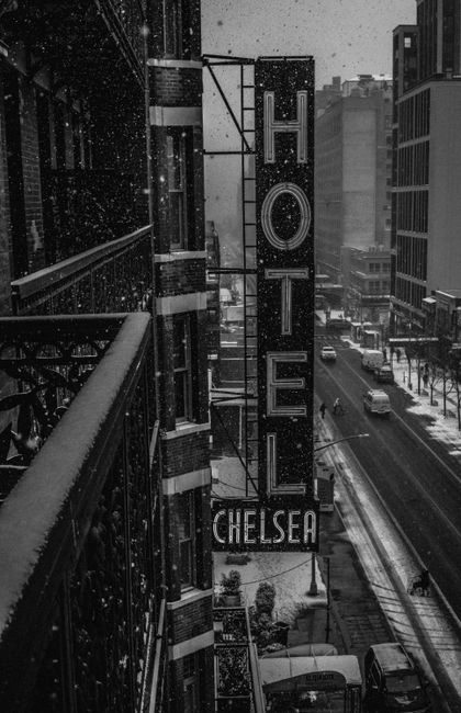 A black and white photograph taken from an upper-floor balcony showing a snowy urban street scene in Chelsea, London. Prominently visible is a vertical hotel sign attached to a building facade, with the large word 'HOTEL' illuminated and the smaller word 'CHELSEA' at the bottom: the sign is suspended by metal brackets. Snow is falling gently, covering the rooftops, street, and parked vehicles below. The street below features moving cars, pedestrians in winter clothing, and a tram track running parallel to the sidewalk. The scene highlights the busy, historic atmosphere of Chelsea, with tall modern buildings lining the street and a sense of active city life despite the snow. This urban setting can be associated with house removals and relocation services, such as those provided by Removal Van Chelsea, especially in the context of moving logistics and furniture transport within the Chelsea area.