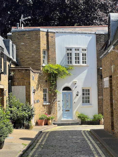 A cobbled residential street in Chelsea featuring narrow pavements lined with various potted plants and small trees in containers on both sides. On the left, there are multi-storey houses with light-c